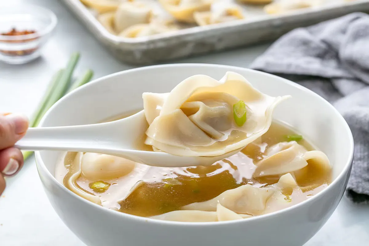 Wonton soup in white bowl, a wonton lifted on a spoon with green onion garnish at One More Bite Dumpling, a Chinese Restaurant in Yucaipa
